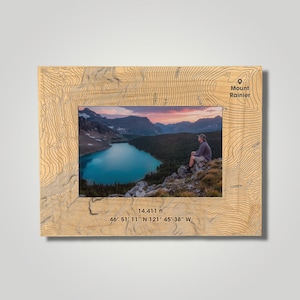 May include: A wooden photo frame with a topographic map design featuring Mount Rainier, Washington. The frame includes the elevation of Mount Rainier, 14,411 feet, and its coordinates, 46° 51' 11" N 121° 45' 38" W. The frame has a photo of a person sitting on a mountain overlooking a lake.