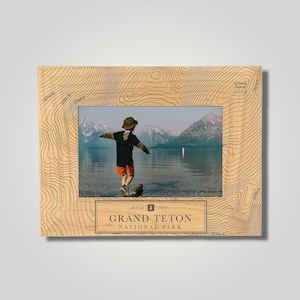 May include: A wooden photo frame with a topographic map design featuring the Grand Teton National Park logo. The frame has a photo of a child standing on a log in a lake with mountains in the background.