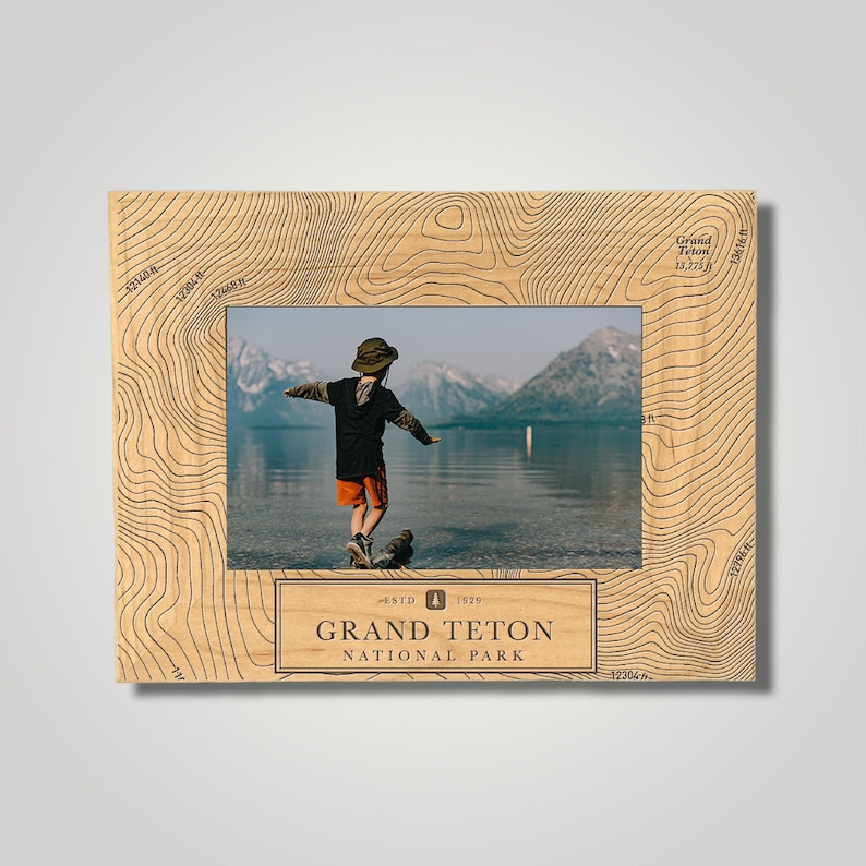 May include: A wooden photo frame with a topographic map design featuring the Grand Teton National Park logo. The frame has a photo of a child standing on a log in a lake with mountains in the background.
