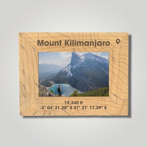 May include: Wooden picture frame with a landscape photo of Mount Kilimanjaro. The frame is engraved with contour lines and the text "Mount Kilimanjaro", along with the altitude of 19,340 ft and coordinates.