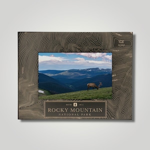 May include: A framed photograph of the Rocky Mountain National Park. The frame is dark brown with a topographic map design. The photograph shows a scenic view of mountains, a blue sky, and an elk grazing on a grassy hillside. The text "Rocky Mountain National Park" is displayed.
