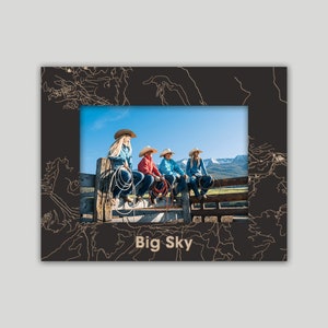 May include: A black picture frame with a gold outline and the text "Big Sky" at the bottom. The frame contains a photo of four people wearing cowboy hats sitting on a wooden fence.
