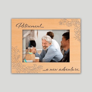 May include: Wooden picture frame with the words "Retirement... ...a new adventure." The frame has a light wood finish and floral corner designs. The frame holds a photo of a smiling man with gray hair.