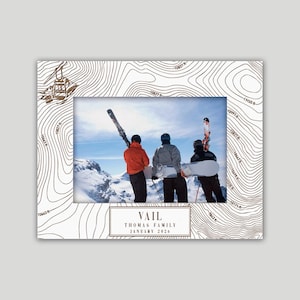 May include: A white photo frame with a topographic map design and a picture of three people with skis and snowboards on a snowy mountain. The frame includes the text "VAIL THOMAS FAMILY JANUARY 2026".