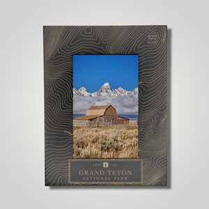 May include: A black picture frame with a topographic map design features a photo of a wooden barn in front of snow-capped mountains. The frame has the text "Grand Teton National Park" and "Est. 1929" on the bottom.
