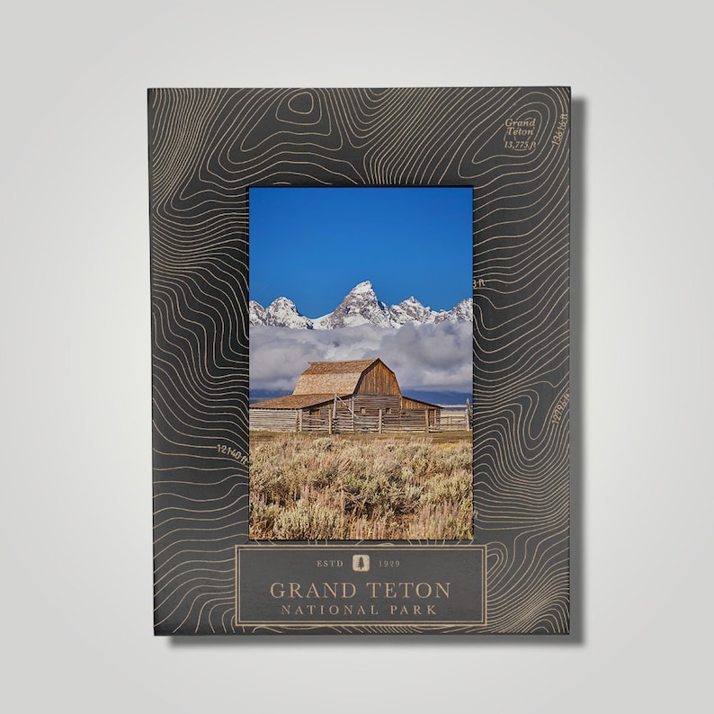 May include: A black picture frame with a topographic map design features a photo of a wooden barn in front of snow-capped mountains. The frame has the text "Grand Teton National Park" and "Est. 1929" on the bottom.