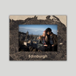 May include: A photo frame featuring a map of Edinburgh, Scotland. The frame is made of wood and has a black finish. The map is printed in white and shows the city's streets and landmarks. The frame includes a photo of a couple sitting on a hill overlooking the city.