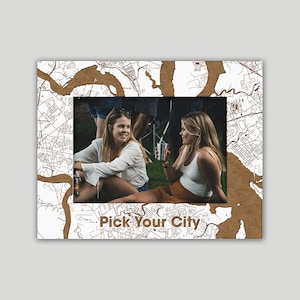 May include: Two friends are sitting on a grassy lawn, smiling and talking. They are holding cups and a can of beverage. The image is framed by a brown map with the text "Pick Your City" at the bottom.