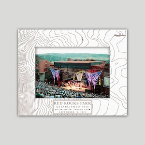 May include: A white picture frame with a topographic map design. The frame holds a photo of a concert at Red Rocks Park. Text on the frame reads "RED ROCKS PARK ESTABLISHED 1936 BILLIE EILISH WORLD TOUR SEPTEMBER 15, 2025."