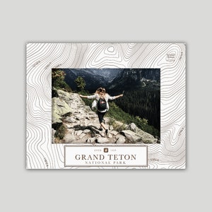 May include: A white and brown topographic map design with a photo of a person hiking in a mountain landscape. The text "ESTD 1929 GRAND TETON NATIONAL PARK" is in brown text on a white background.