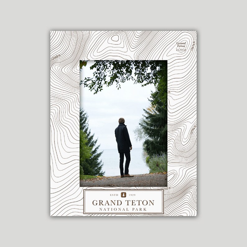 May include: A white photo frame with a brown topographic map design. The frame has a cutout for a photo and a brown label that reads "ESTD 1929 GRAND TETON NATIONAL PARK".