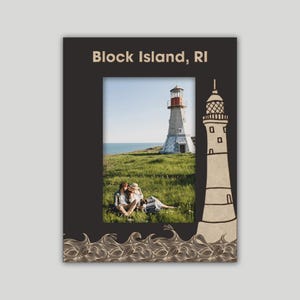 May include: A black picture frame with a white lighthouse and a couple sitting on a grassy hill in the background. The frame has a brown lighthouse illustration and a wave pattern at the bottom. The text "Block Island, RI" is at the top of the frame.