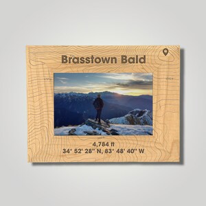 May include: A wooden photo frame with a topographic map design features a photo of a person standing on a mountaintop with a view of a snowy mountain range. The frame includes the text "Brasstown Bald", "4,784 ft", and "34° 52' 28" N, 83° 48' 40" W".
