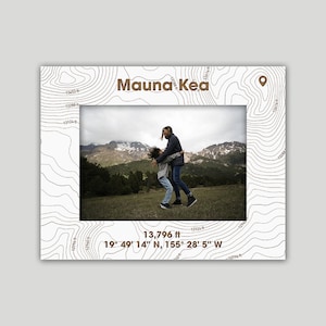 May include: A photo of two people standing in a field with mountains in the background. The image is framed with a topographic map design and the text "Mauna Kea" and "13,796 ft 19° 49' 14" N, 155° 28' 5" W".