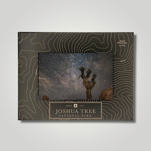 May include: A dark brown wooden frame with a gold topographic map design. The frame has a photo insert of a Joshua Tree National Park night sky with a Joshua Tree in the foreground. The text "ESTD 1994 JOSHUA TREE NATIONAL PARK" is printed on the frame.