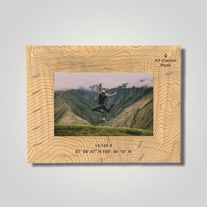 May include: Wooden picture frame with a topographic map design. The frame surrounds a photo of a person jumping in front of a mountain range. The frame includes the text "Kit Carson Peak" and coordinates, with an elevation of 14,165 ft.