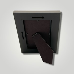 May include: Black picture frame with a brown cardboard backing and a fold-out easel stand on the back.