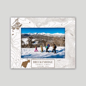 May include: A white photo frame with a topographic map design in brown. The frame holds a photo of a family skiing in the mountains. The text "BRECKENRIDGE THOMAS FAMILY JANUARY 2026" is printed on the frame.