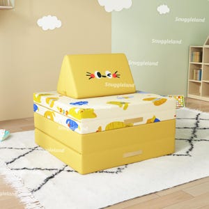 May include: A yellow and white children's play sofa with a cat face on the back. The sofa is made of foam and has a removable cover. The sofa is on a white rug with a black and white pattern.