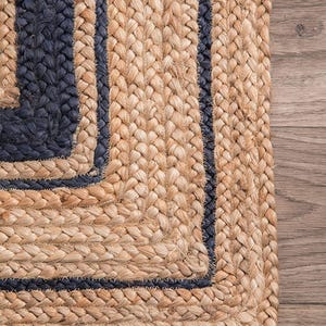 Puede incluir: Primer plano de una alfombra de yute natural con un diseño tejido. La alfombra presenta un color marrón claro con bordes y rayas azul oscuro, creando un patrón contrastante. La textura es visible, mostrando las fibras trenzadas.