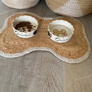 Puede incluir: Dos cuencos de cerámica blanca para perros con huellas de patas negras y el texto "The Greenbrier" en una alfombra de yute marrón en forma de hueso.