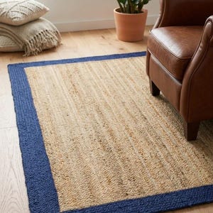 Puede incluir: Una alfombra rectangular con un centro de yute tejido en color tostado natural y un borde grueso de color azul marino sólido. La alfombra presenta una construcción trenzada y texturizada, colocada sobre un suelo de madera clara.