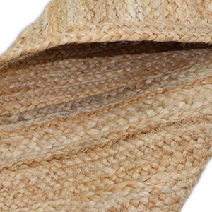 May include: Close-up of a natural jute rug with a braided texture. The rug features varying shades of tan and brown, with visible woven patterns. The edges of the rug are also visible, showing the thickness and construction of the material.