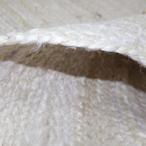 Könnte beinhalten: Eine Nahaufnahme eines gewebten Teppichs aus natürlichen, hellbeigen Fasern. Die Kante des Teppichs weist einen dicken, geflochtenen Rand auf, der einen markanten, erhabenen Abschluss bildet, während der Hauptteil eine texturierte Webstruktur zeigt.