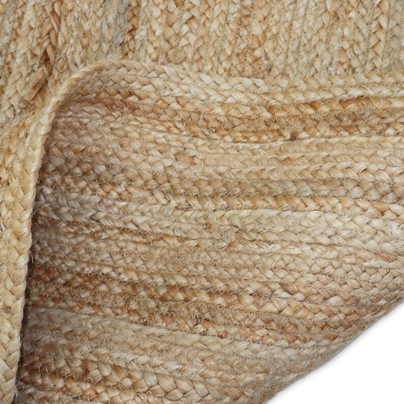 May include: Close-up of a natural jute rug with a woven texture. The rug is a light tan color with visible fibers and a rolled-up edge, showcasing the detailed craftsmanship and organic material.