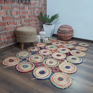 May include: A colorful jute rug made of woven circles in shades of brown, red, blue, green, and yellow. The rug is laid out on a wooden floor and features a round jute pouf and a white planter with a green plant.