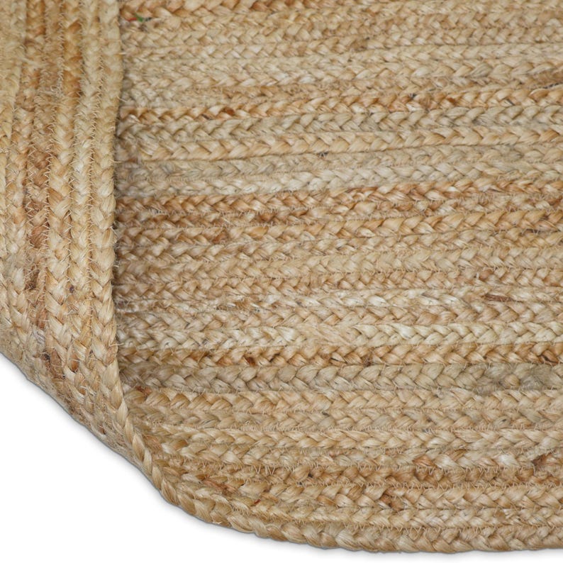 May include: Close-up of a natural fiber rug with a braided texture. The rug is made of light brown jute fibers, with a rolled-up edge revealing the underside. The weave creates a textured surface, suitable for home decor.