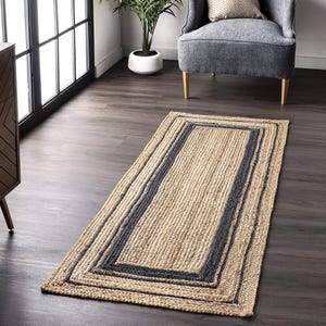 Puede incluir: Una alfombra rectangular hecha de fibras naturales trenzadas de color tostado, con un borde prominente de color gris carbón oscuro que enmarca la sección central. La alfombra tiene una apariencia texturizada y trenzada con un patrón uniforme.