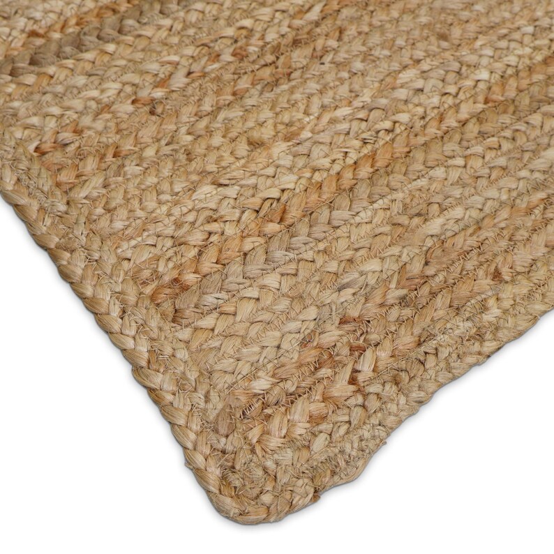 May include: Close-up of a natural jute rug. The rug is made of woven, braided fibers in shades of tan and brown. The texture is visible, with a braided border and a diagonal pattern across the surface. The corner of the rug is visible against a white background.