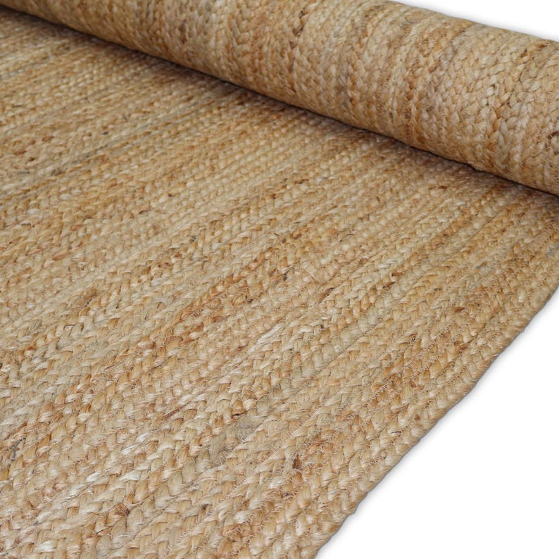 May include: A rolled-up, natural-toned jute rug. The rug is made of woven fibers, with a visible braided pattern. The color is a warm, light brown, and the texture appears coarse. The rug is partially rolled, revealing the woven construction.