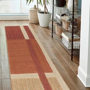 May include: A long, rectangular, geometric patterned rug in shades of beige, brown, and red. The rug is made of natural fibers and has a woven texture.