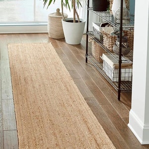 May include: A rectangular, natural-toned jute runner rug. The rug is placed on a hardwood floor, with a woven texture and a neutral color. The rug is long and narrow, suitable for hallways or entryways. The rug is surrounded by plants and decorative items.