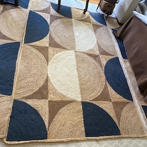 Pode incluir: Um tapete retangular grande com um padrão geométrico. O tapete apresenta um design repetitivo de círculos e quartos de círculo em tons de bege, castanho e azul marinho. O tapete é feito de um material de fibra natural.