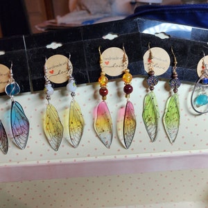 May include: A set of eight pairs of handmade earrings featuring various designs. The earrings are displayed on a card with the text "Handmade with love". The earrings feature a variety of colours, including blue, green, yellow, and red. Some earrings feature a dragonfly wing design, while others feature a teardrop design.