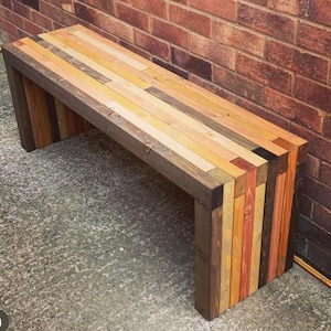 May include: A rustic wooden bench crafted from reclaimed wood planks. The bench has a dark brown stain and is made up of various shades of wood, including light brown, yellow, and red. The bench is sitting on a concrete surface.