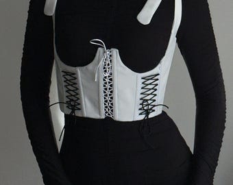 May include: A white corset top with black lacing and white tie straps. The corset has a structured design with a curved neckline and is worn over a black long-sleeved top and black skirt.