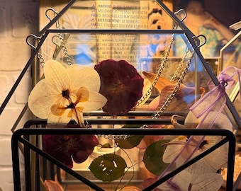 May include: A geometric glass terrarium with a black metal frame, containing dried flowers in white, purple, and burgundy. The terrarium is suspended by chains. The text on the glass reads: "WARNING: GLASS IS FRAGILE..." The measurements are in centimeters.