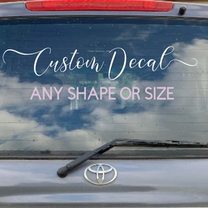 May include: A white custom decal on the back window of a silver car. The decal says "Custom Decal" in a cursive font and "ANY SHAPE OR SIZE" in a smaller font.