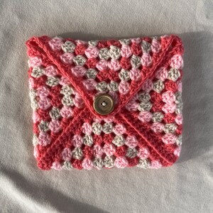 May include: A pink and beige crocheted envelope clutch with a wooden button closure.