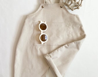Könnte beinhalten: Eine beige Cordhose mit Latz und einer großen Tasche auf der Vorderseite. Ein Paar weiße Sonnenbrillen ist in die Tasche gesteckt.