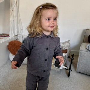 May include: A young child wearing a gray corduroy shirt and matching pants. The child is standing in a room with a white wall and a gray carpet.