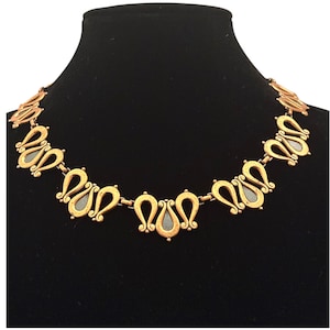 May include: Gold-toned necklace with a series of stylized teardrop-shaped links. Each link features a dark gray center and ornate detailing. The necklace is displayed on a black background.
