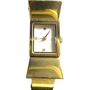 Puede incluir: Reloj de pulsera dorado con esfera rectangular blanca. El reloj presenta un logotipo de pica y tres pequeños diamantes. La correa tiene un diseño en forma de lazo. El reloj mide aproximadamente 2,5 cm de ancho.