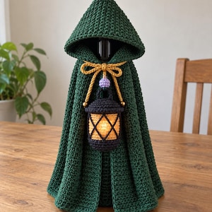 Puede incluir: Una capa con capucha de ganchillo verde oscuro drapeada sobre una botella. La capa presenta un cordón amarillo con una pequeña cuenta de color lavanda y un adorno de linterna de ganchillo negro con un centro naranja brillante.