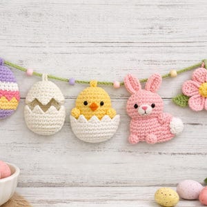 May include: A handmade Easter garland featuring crocheted decorations. The garland includes a purple and yellow egg, a cracked egg, a yellow chick, a pink bunny, and a pink flower. The decorations are strung on a green cord with wooden beads.