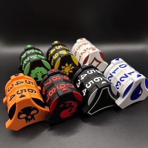 Könnte beinhalten: Eine Sammlung von sieben vielseitigen Würfeln in verschiedenen Farben, darunter Orange, Schwarz, Weiß und Grün. Jeder Würfel hat ein anderes Symbol und eine andere Zahlenanordnung, mit Zahlen von 0 bis 9. Die Würfel sind für Tabletop-Spiele konzipiert.